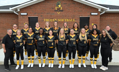 Team photo of the softball team in black and gold uniforms standing in front of the brick fieldhouse.