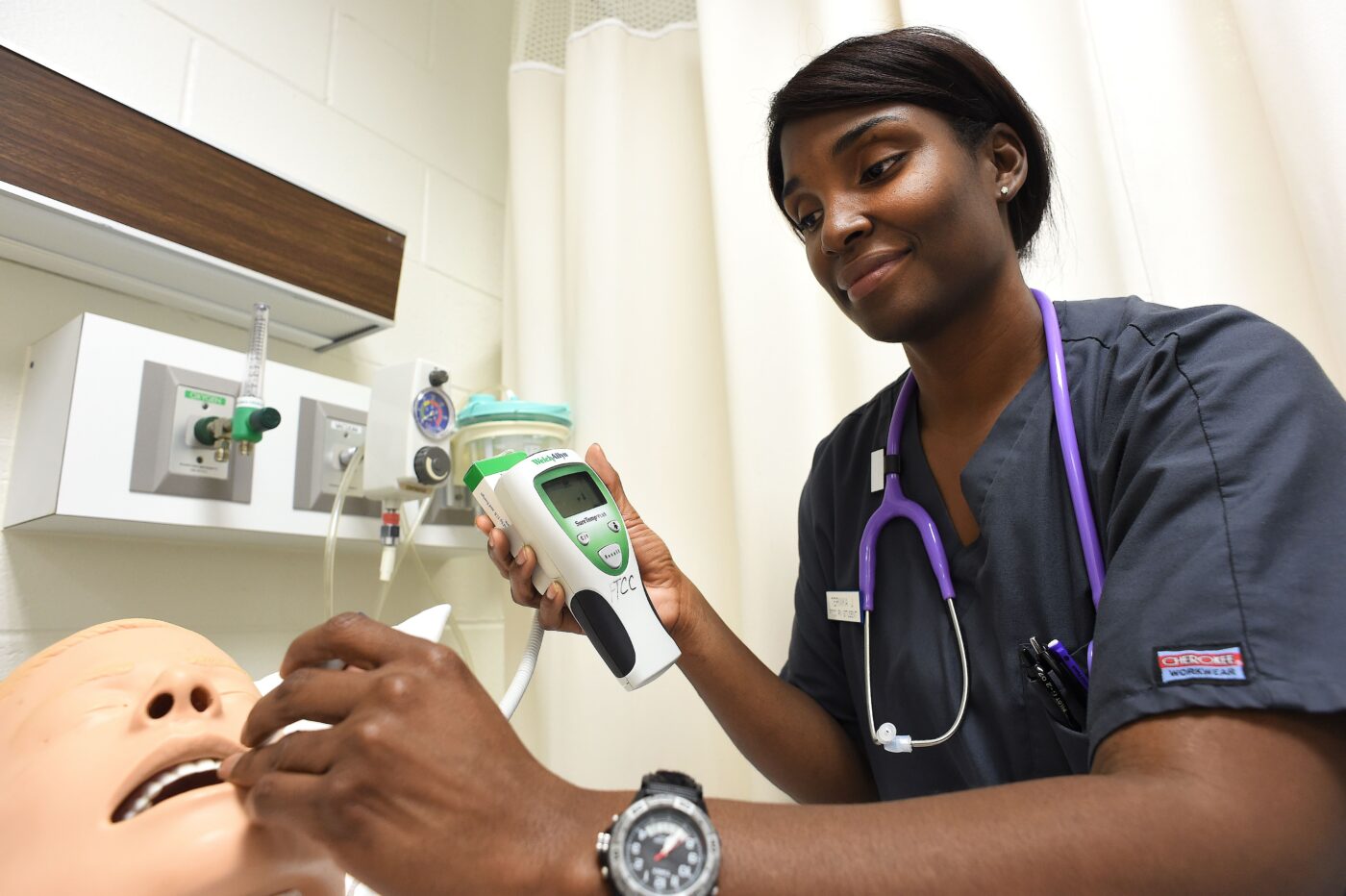 The image shows a medical professional, possibly a nurse, using a medical device on a mannequin in a clinical setting. The nurse is wearing a grey scrub top with a purple stethoscope around her neck. She is holding a device that seems to be used for checking vital signs, such as a thermometer or a pulse oximeter, and appears to be performing a procedure on the mannequin, which is part of a training exercise. The background includes medical equipment and a hospital curtain.