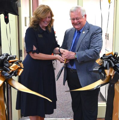 Two people use oversized scissors to cut a ribbon in front of a door