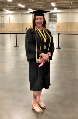 A graduate stands slights turned to the left. She has her hands clasped in front of her and is wearing a gold cord and a medal on a black and gold ribbon over her gown. 