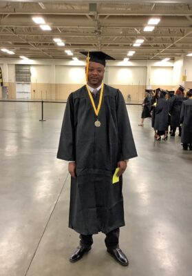 A man wearing a graduation cap and gown stands facing the camera. He is wearing a medal on a black and gold ribbon around his neck and is holding a green piece of paper in his hand.