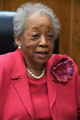 A close-up photo of Marye Jeffries wearing a pink blouse and blazer with a large pink flower on the lapel. She is wearing pearl earrings and necklace.