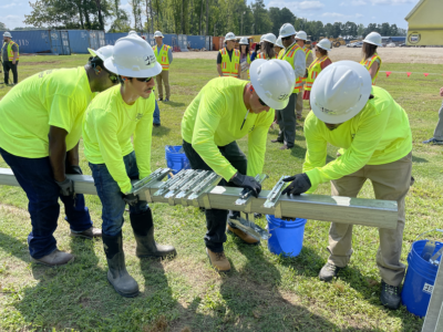 Four men in hard hats and high visibility shirts lift a beam in a field near a solar farm.
