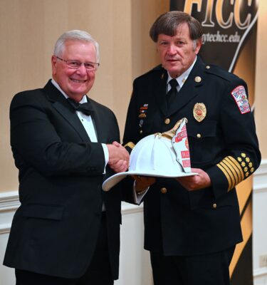 Larry Keen and Freddy Johnson shake hands while holding a white fire chief's hat.