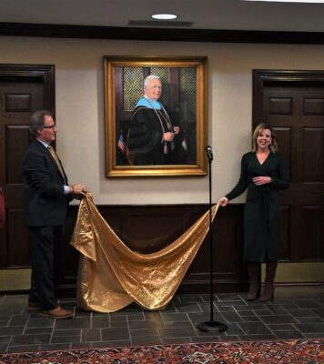 Dr Mark Sorrells and Robin Deaver stand on either side of an oil painting portrait of Dr. Larry Keen. They are hold the ends of a gold cloth that had been covering the painting.