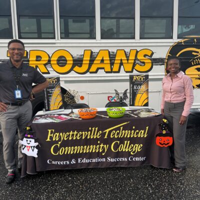 Marvin Price and Tiffany Lesane stand beside a table in front of a FTCC Trojans bus.
