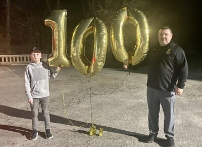 A boy and a man stand on each side of three balloons that form the number 100.