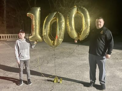 A boy and a man stand on each side of three balloons that form the number 100.