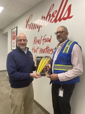 Two men hold an FTCC folder between them. They are standing in a well-lit hallway and looking at the camera.