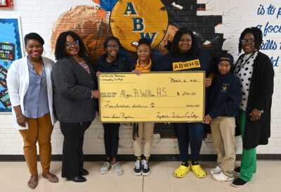 A group of women and girls stand in front of a mural on a painted brick wall. The group is holding a large ceremonial check for $500 made out to Alger B Wilkins HS from Fayetteville Technical Community College.