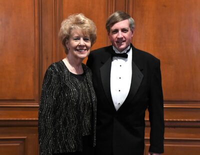 Carol and Bill Conklin dressed in formal wear stand in front of a wood-paneled wall.