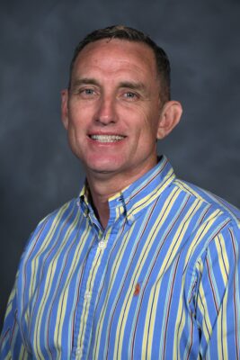 A studio portrait of Scot McCosh wearing a blue and yellow striped button up shirt.
