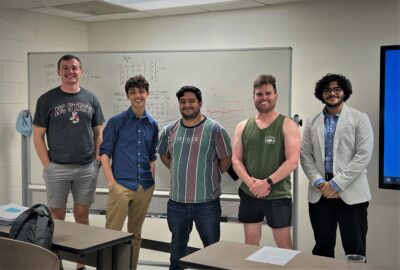 A group of males students stand in a line at the front of a classroom. A whiteboard with algebra notes written on it is visible behind them.