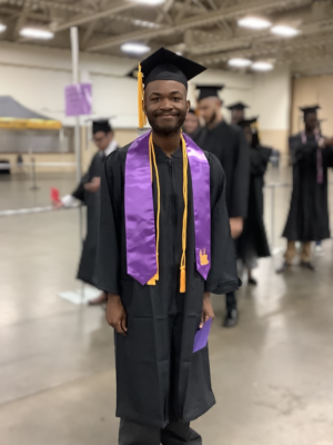 Alexander Johnson, a graduate wearing a cap, gown, gold cord and purple stole, stands in a large staging area.