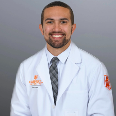 Brandon Little, with close-cropped dark hair and beard, wears a collar shirt, necktie and white pharmacist's coat.