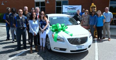 A group of people stand around a white car with a large green ribbon on the hood.