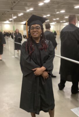 Nakita Scott, dressed in a graduation cap and gown, stands with her arms crossed at the wrists in the staging area before graduation.