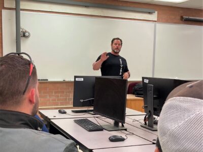 Andrew Ruhland stands at the front of a room speaking. There are computer monitors and students, seen from behind, in the foreground of the photo.