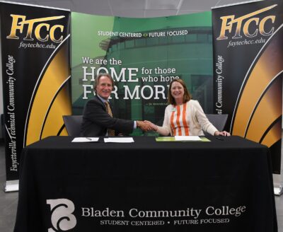 Mark Sorrells and Amanda Lee sit behind a table and shake hands while looking at the camera. There are backdrops behind them with FTCC and Bladen Community College logos on them.