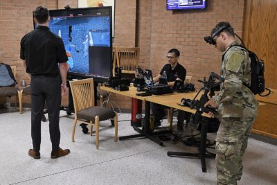 A man in a military uniform wears a weapons simulator system. Another man sits behind a table and a third man stands in front of a monitor.