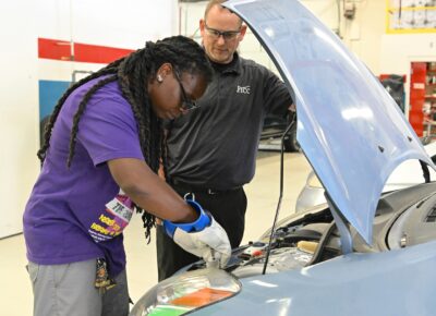 Tiffany Bethea wears protective gloves while she works under the hood of an electric vehicle as instructor Brian Oldham looks on.