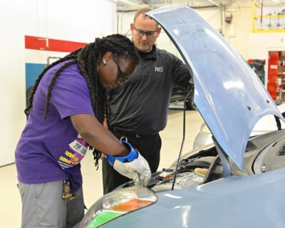 Tiffany Bethea wears protective gloves while she works under the hood of an electric vehicle as instructor Brian Oldham looks on.
