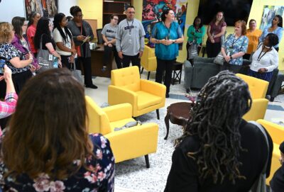 Visitors stand inside the collegiate recovery program center. Bright yellow armchairs are in the center of the photo.