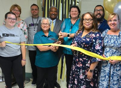 A group of people stand behind a black and yellow ribbon. One woman holds a pair of large scissors prepared to cut the ribbon.