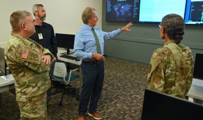 Mark Sorrells stands in the middle of a classroom with Cyber Threat maps on digital displays behind him.