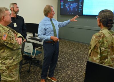 Mark Sorrells stands in the middle of a classroom with Cyber Threat maps on digital displays behind him.