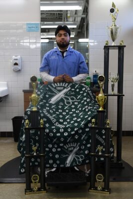Barber student Danny Garcia stands behind a barber's chair with three trophies standing around him.