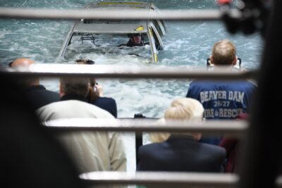 A man in safety gear sits inside a car that is almost entirely submerged in an indoor tank. Spectators can be seen from behind in the foreground of the photo.