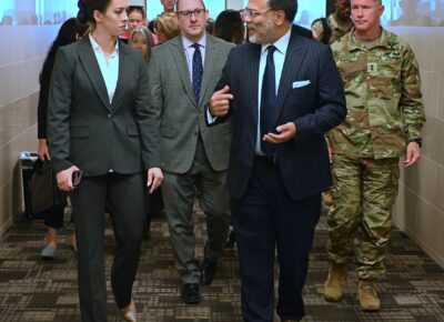 A woman and a man in suits walk at the front of a group of people down a hallway,