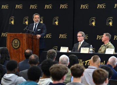 Harry Coker speaks while standing at a podium. Two men are seated at a table next to the podium. The back of the audience is visible in the foreground.