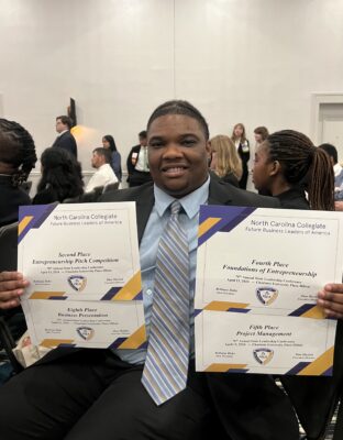 A student in a suit holds up four awards certificates.