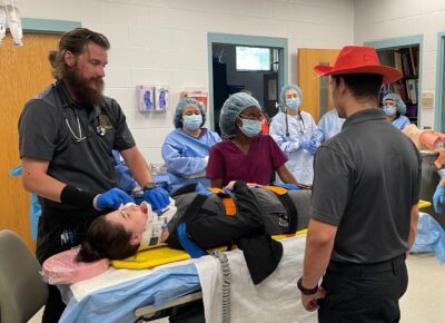 Allied Health students stand around a student pretending to be injured on an operating table in a classroom.