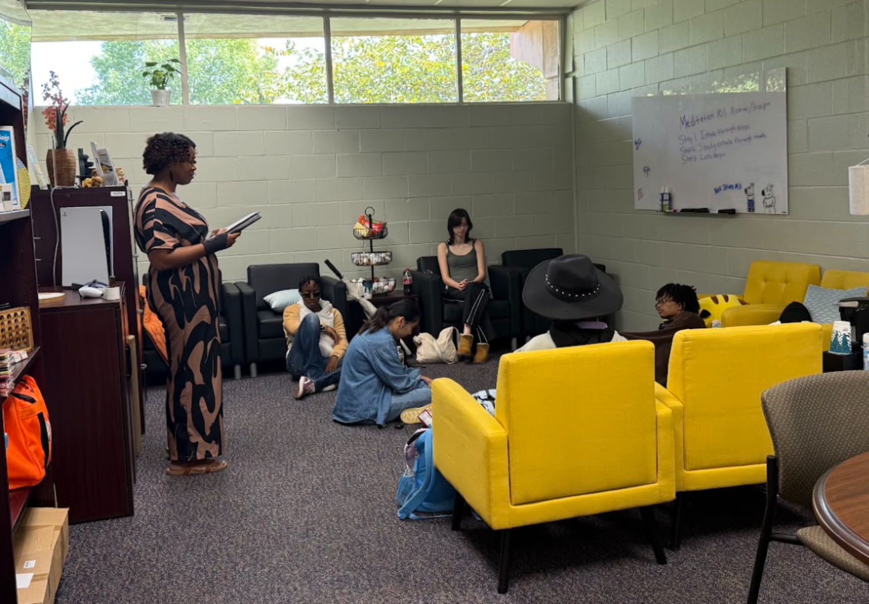 Students Gather In A Lounge With Yellow Chairs As A Facilitator Reads From A Notebook During A Group Session.