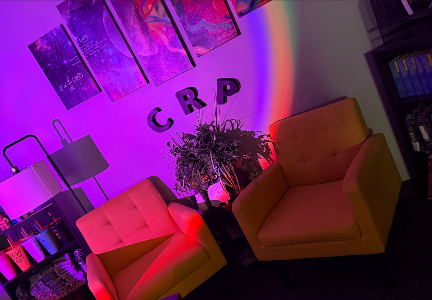 Two Yellow Lounge Chairs With A Plant And Crp Wall Letters, Under Colorful Purple And Rainbow Lighting.