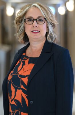 A head-and-shoulders portrait of a woman with blond hair and glasses, dressed professionally.