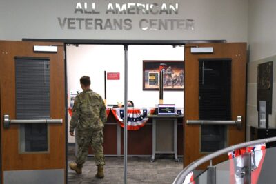 A man in a military uniform walks through double doors under a sign that says All American Veterans Center