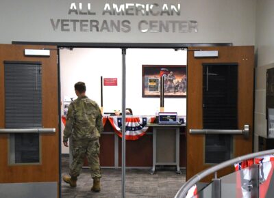 A man in a military uniform walks through double doors under a sign that says All American Veterans Center