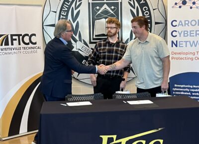 A man in a suit shakes hands with a younger man while another man looks on.