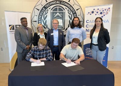 Two men sit at a table and sign papers while a group of people stand behind them.