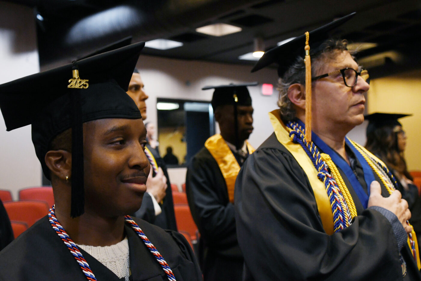 A close-up of two people dressed in graduation caps and gowns, standing with their hands over their hearts.