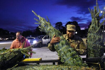 A man in a military uniform lifts a Christmas tree off of a trailer. A second man is in the background also lifting a tree off the trailer.