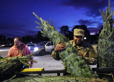 A man in a military uniform lifts a Christmas tree off of a trailer. A second man is in the background also lifting a tree off the trailer.