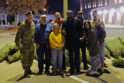 A group of people stand in front of several Christmas trees laying on the ground.