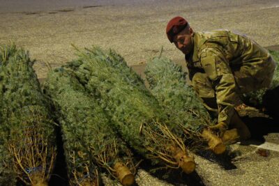 A man in a military uniform bends down to lift a Christmas tree off the ground.