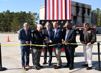 A group of six men cut a ribbon in front a United States flag and a podium built to resemble the front bumper of a fire engine.