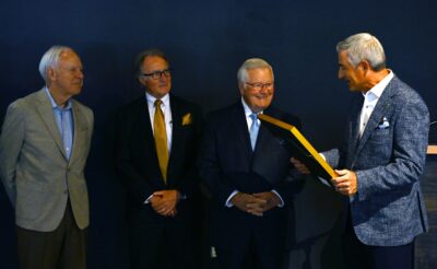 Four men in suits stand in a row. The man on the right holds a gold framed certificate.